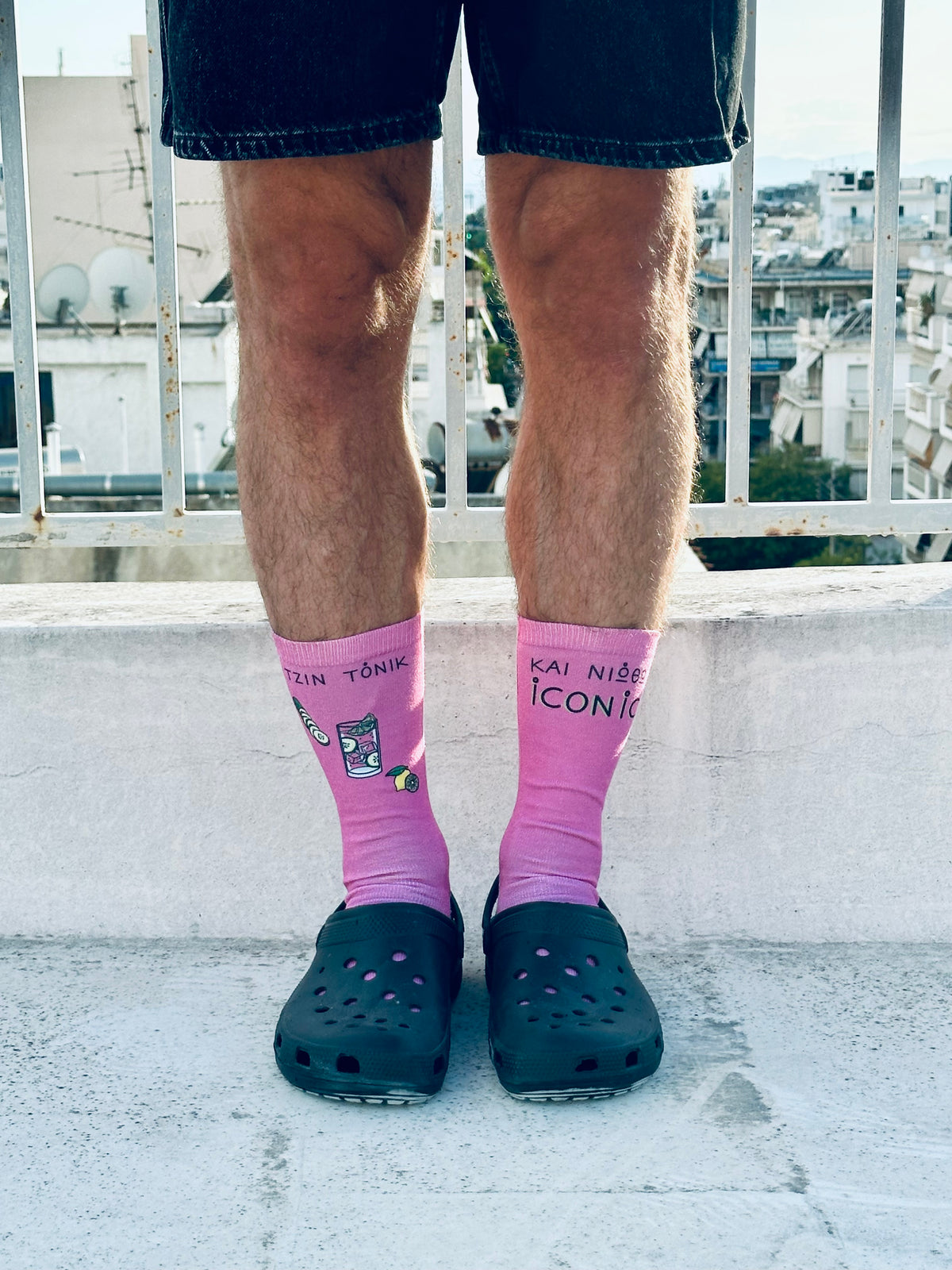 A pair of pink socks with text and illustrations, worn on a person's legs protruding from rolled-up blue jeans, against a backdrop of a cityscape.
