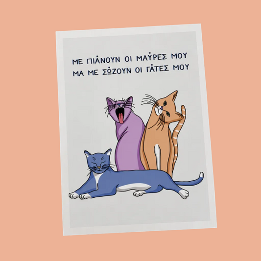 A rectangular greeting card with a white border lies on a wooden tabletop. The card features three cartoon cats in purple, orange, and blue, with Greek text above them.