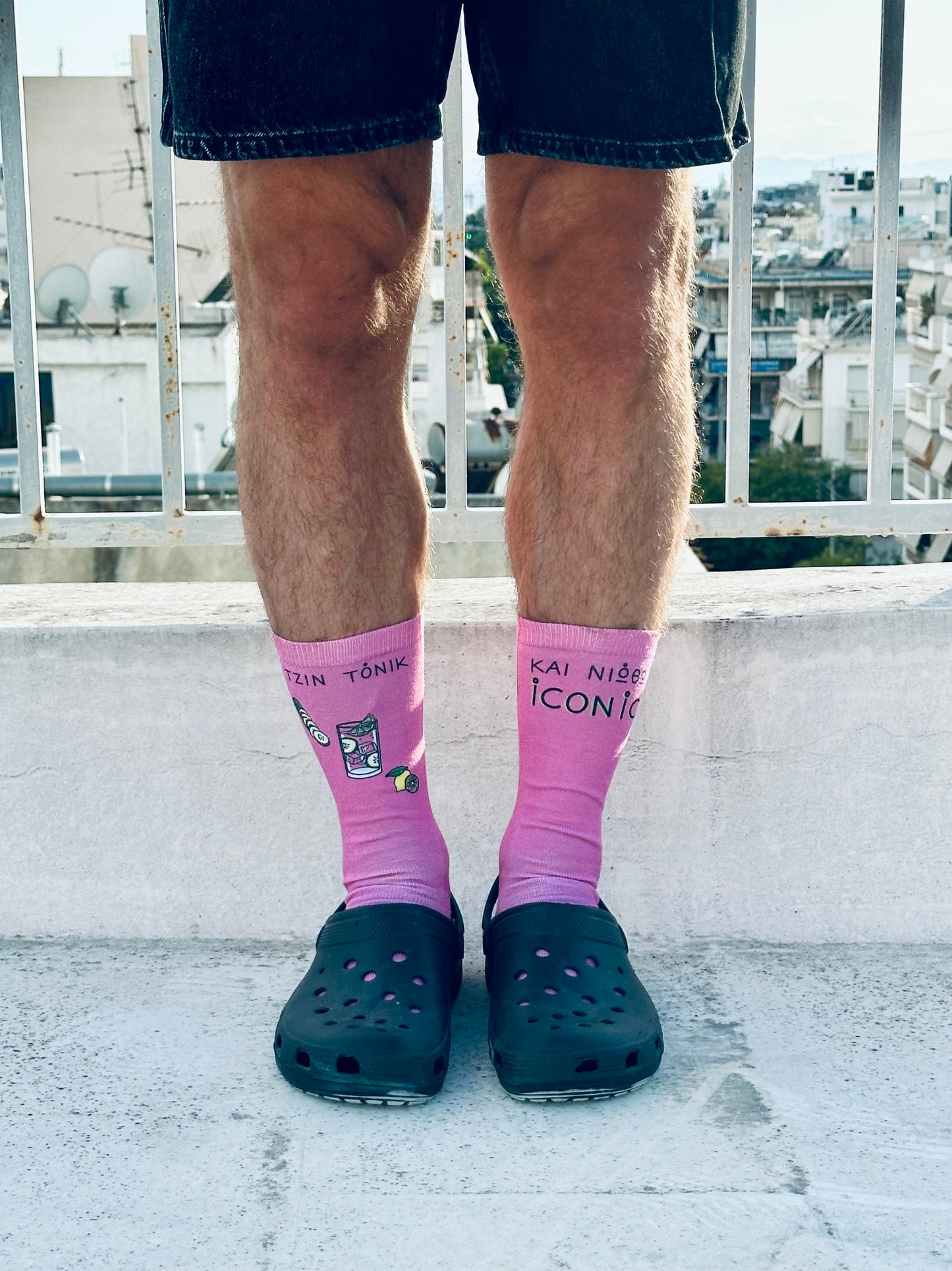 A pair of pink socks with text and illustrations, worn on a person's legs protruding from rolled-up blue jeans, against a backdrop of a cityscape.