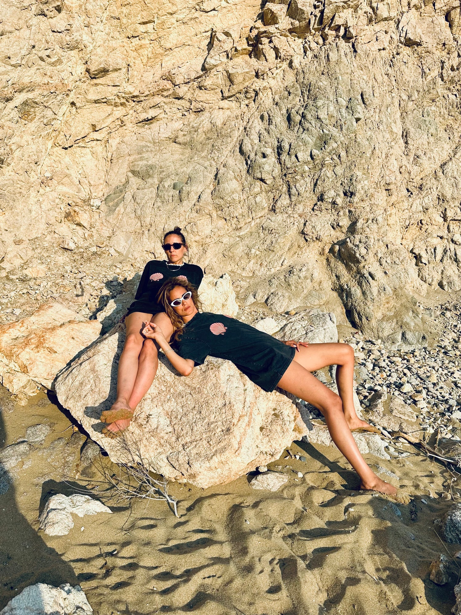 Two people lounging on a rock at the beach with rocky cliffs in the background.