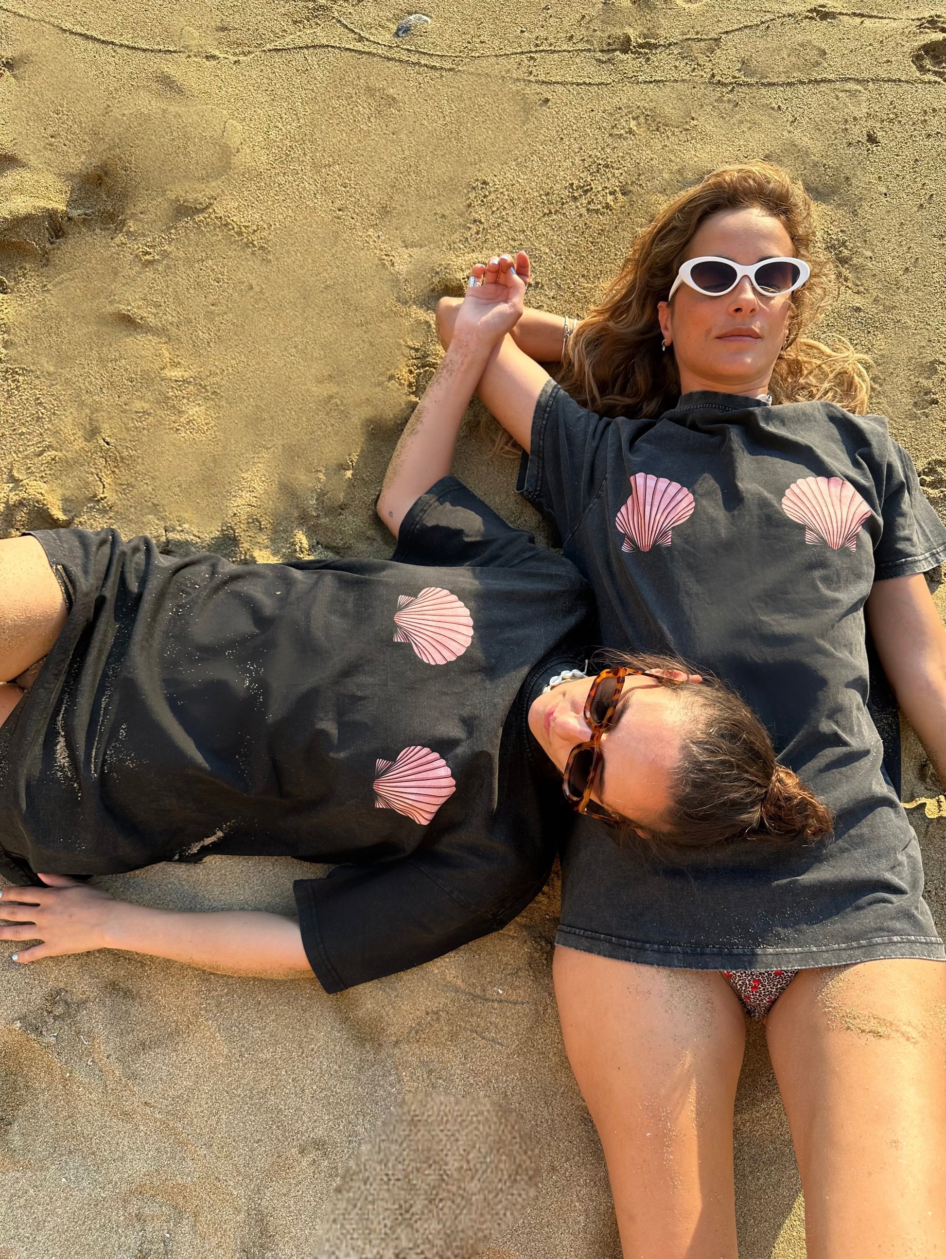 Two people lying on sand wearing matching black dresses with pink shell patterns.