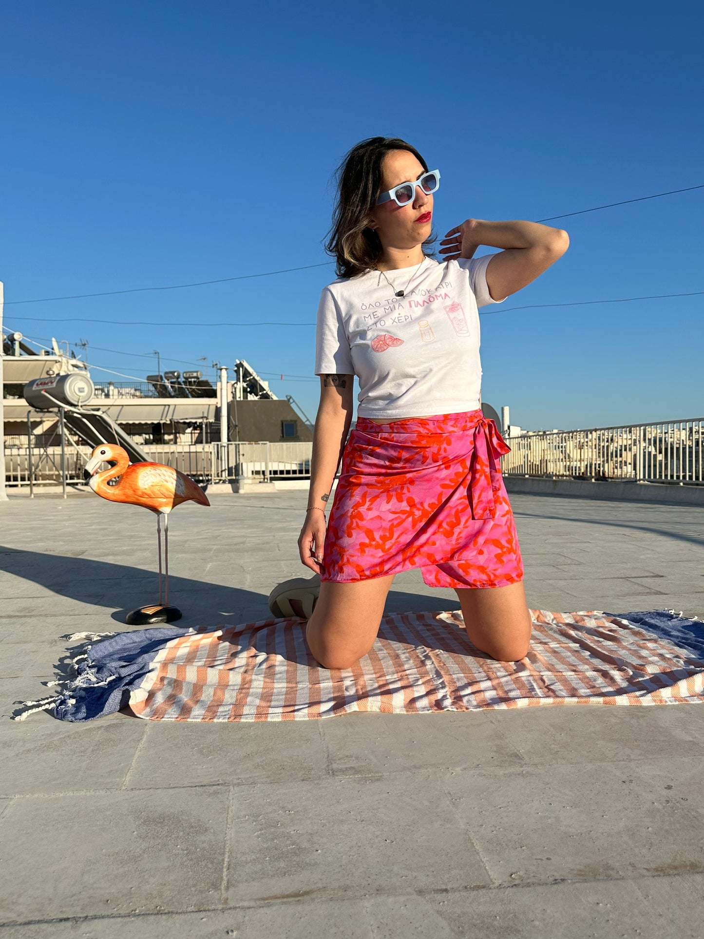 Woman in a white t-shirt and pink skirt standing on a rooftop with a flamingo decoration and blue sky.