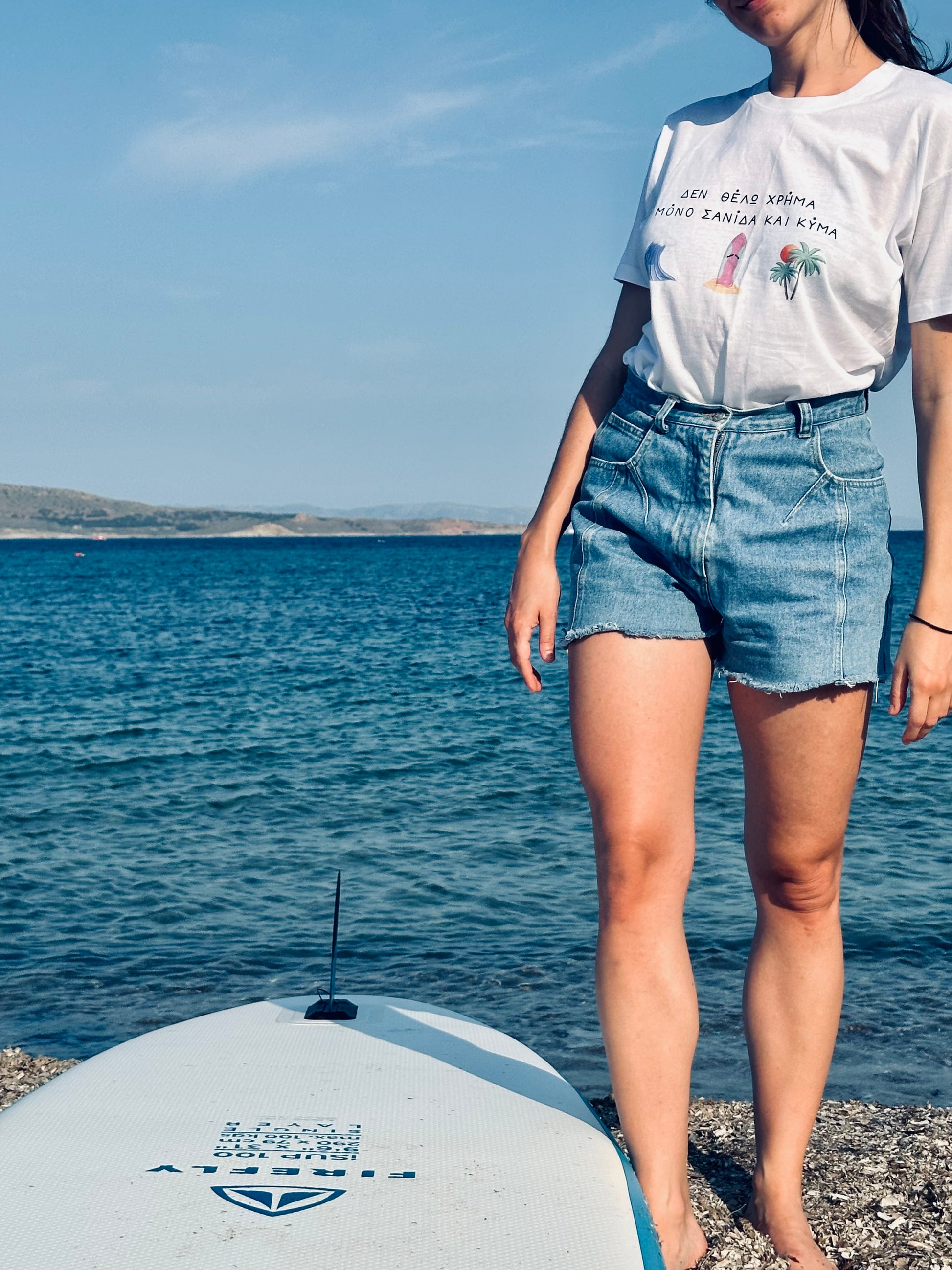 Person standing on a beach with a paddleboard, wearing a white t-shirt and denim shorts.