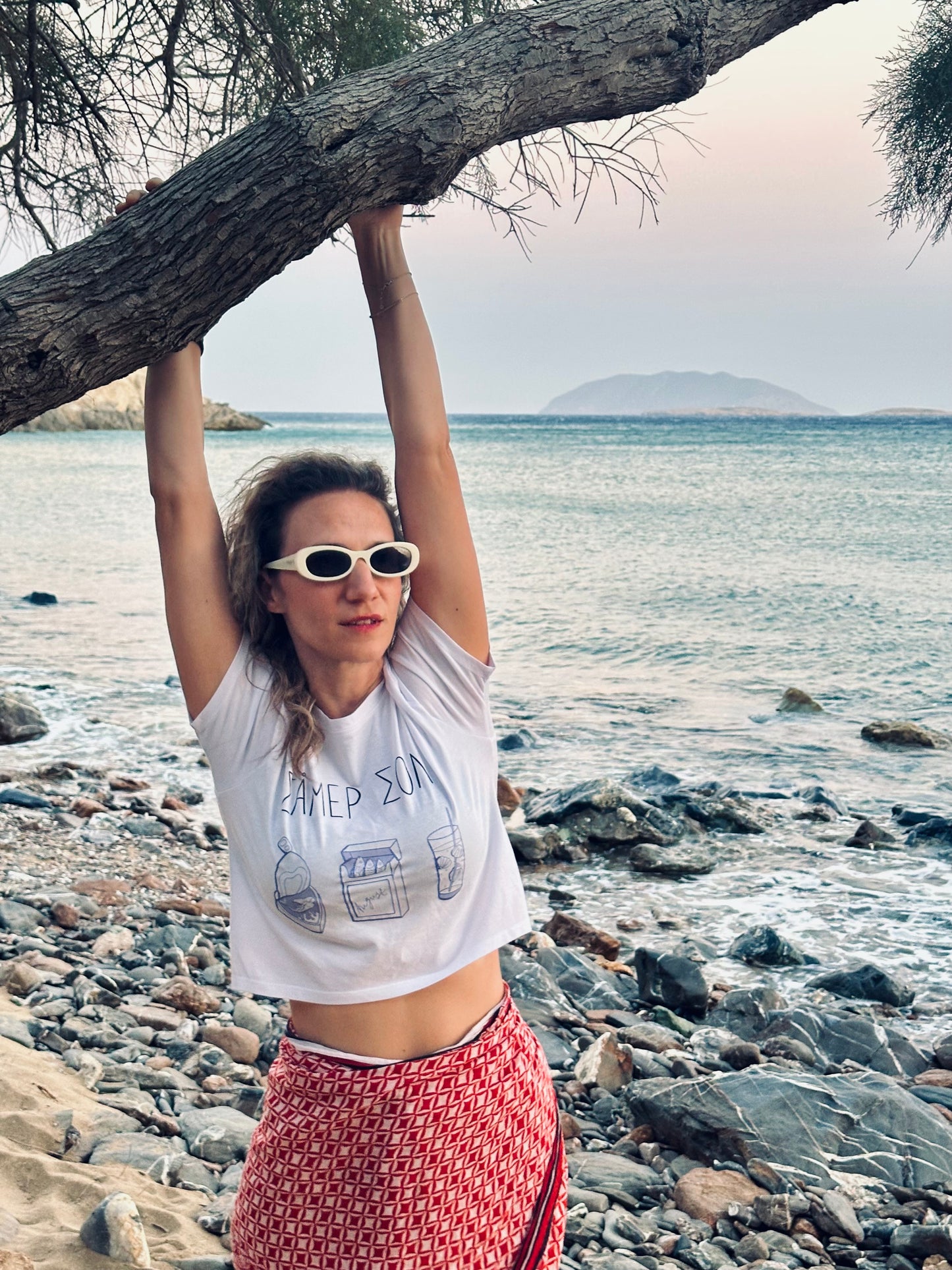 Woman on a rocky beach with ocean view, wearing sunglasses and a white t-shirt.
