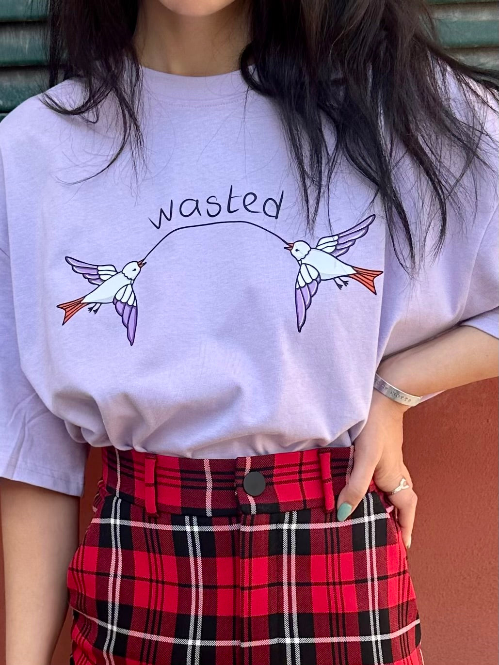 Person wearing a light purple t-shirt with a graphic design and text, paired with a red plaid skirt.