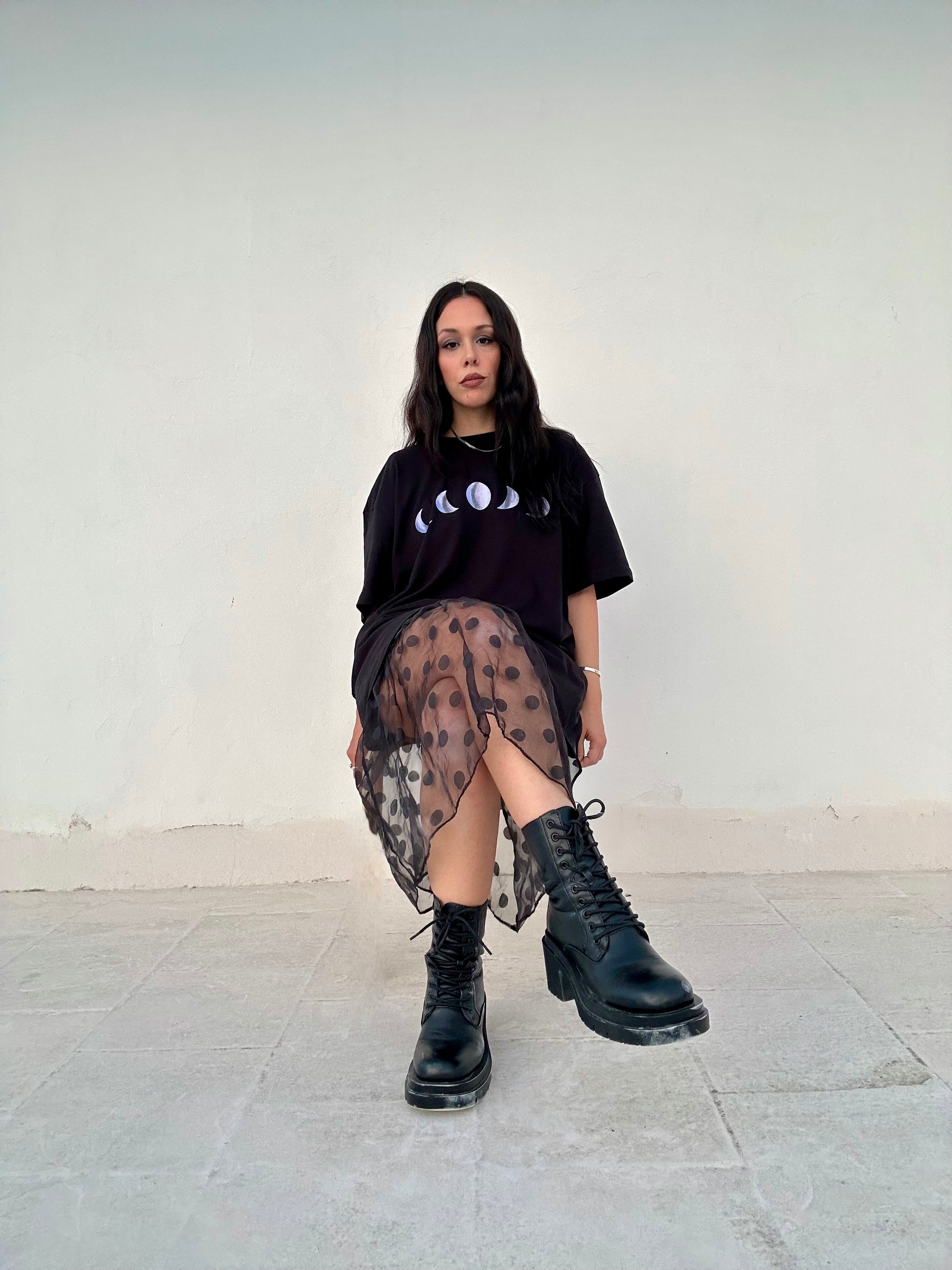 Person wearing a black t-shirt with graphic design, sheer patterned skirt, and black boots against a white wall.