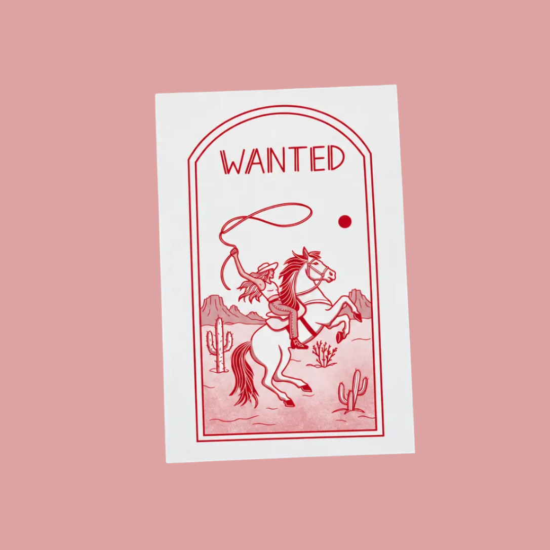 A white rectangular card with a red western-style 'WANTED' illustration of a rider on a horse, placed on a light wood grain surface.