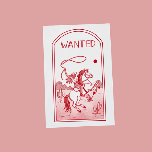 A white rectangular card with a red western-style 'WANTED' illustration of a rider on a horse, placed on a light wood grain surface.